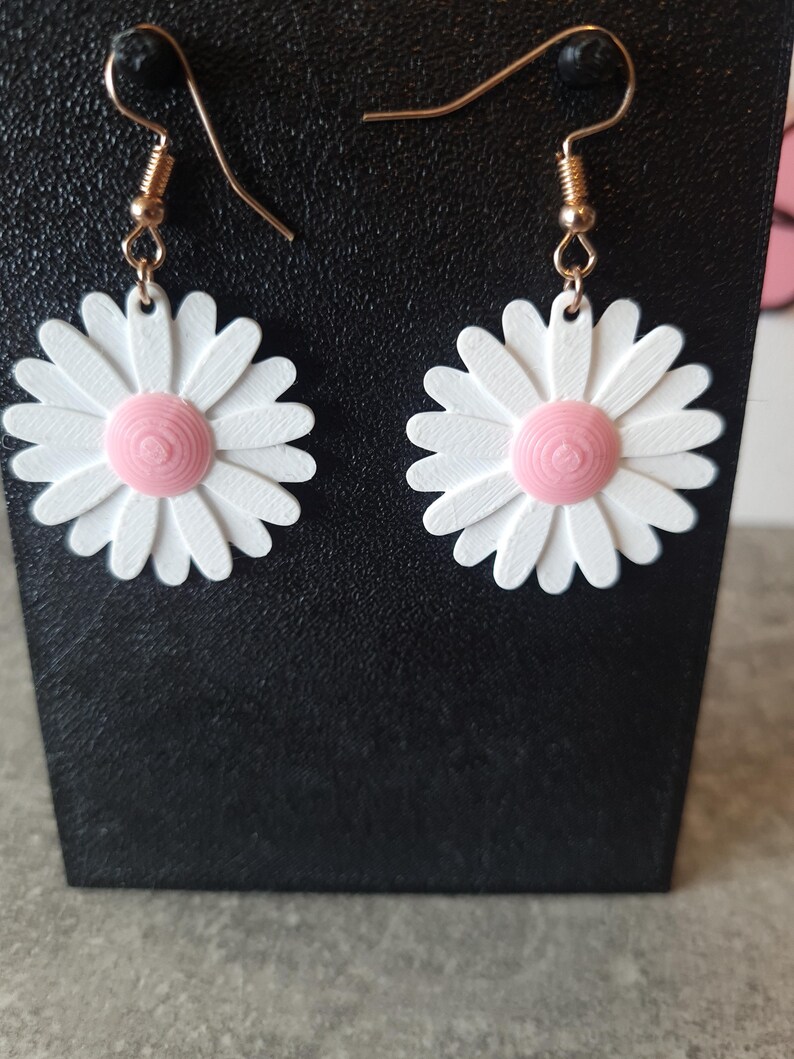 May include: A pair of daisy earrings. Each earring features a white daisy with a pink center. The daisy petals are detailed. The earrings have gold-colored hooks.