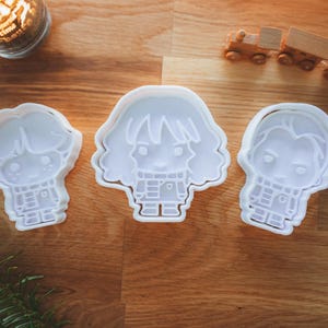 May include: Three white plastic cookie cutters in the shape of cartoon characters, arranged on a wooden surface. The cutters depict a boy, a girl, and another boy. Decorative candles and a toy train are in the background.