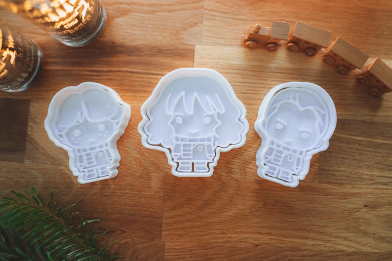 May include: Three white plastic cookie cutters in the shape of cartoon characters, arranged on a wooden surface. The cutters depict a boy, a girl, and another boy. Decorative candles and a toy train are in the background.
