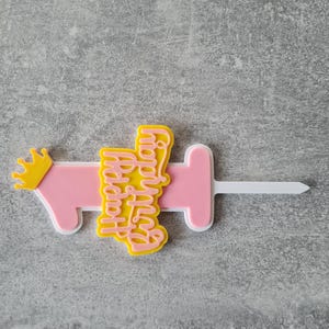 Op de afbeelding: Een roze en gele taarttopper met de tekst "Happy Birthday" in een scriptlettertype. De topper heeft de vorm van een spuit met een gele kroon aan de linkerkant. De topper is gemaakt van plastic.