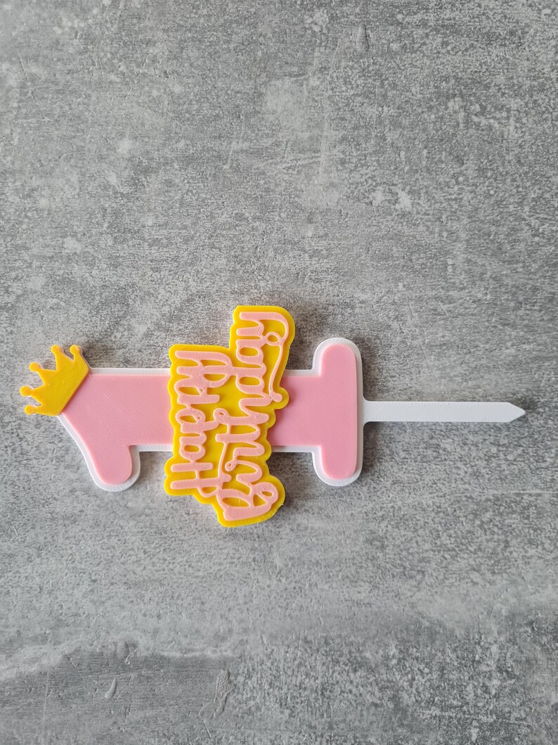 Op de afbeelding: Een roze en gele taarttopper met de tekst "Happy Birthday" in een scriptlettertype. De topper heeft de vorm van een spuit met een gele kroon aan de linkerkant. De topper is gemaakt van plastic.