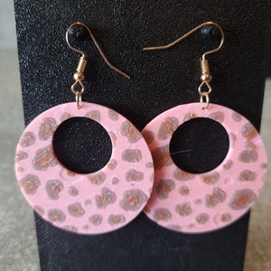 May include: Pink hoop earrings with a leopard print pattern in shades of brown and gray. The earrings have gold-colored ear wires and are displayed on a black stand.