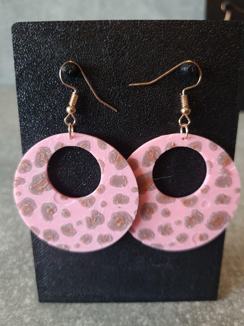 May include: Pink hoop earrings with a leopard print pattern in shades of brown and gray. The earrings have gold-colored ear wires and are displayed on a black stand.