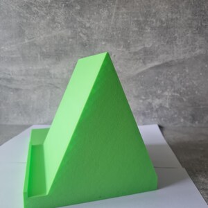 May include: A bright green, triangular phone stand. The stand has a flat base with a small ledge to hold a phone or tablet. The stand is made of a solid, textured material and is designed to prop up a device for hands-free viewing.