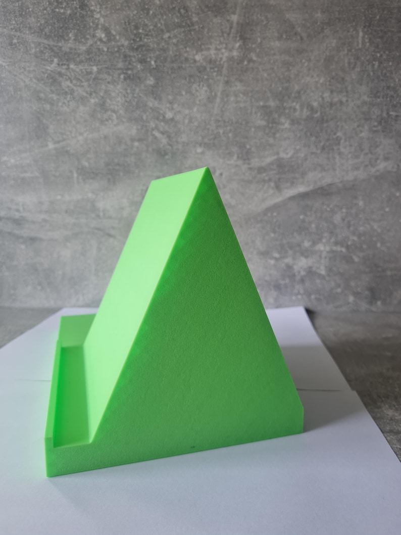 May include: A bright green, triangular phone stand. The stand has a flat base with a small ledge to hold a phone or tablet. The stand is made of a solid, textured material and is designed to prop up a device for hands-free viewing.