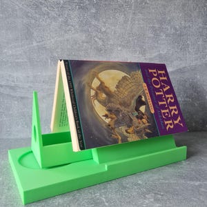 May include: A bright green book stand holding a copy of "Harry Potter and the Prisoner of Azkaban" by J.K. Rowling. The book has a purple cover with an illustration of a creature. The stand is made of a solid, smooth material.