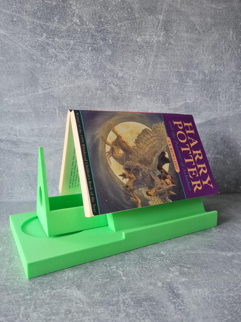 May include: A bright green book stand holding a copy of "Harry Potter and the Prisoner of Azkaban" by J.K. Rowling. The book has a purple cover with an illustration of a creature. The stand is made of a solid, smooth material.