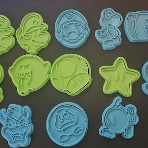 May include: A selection of green and blue plastic cookie cutters depicting characters from a popular video game. The set includes Mario, Luigi, Yoshi, a Bullet Bill, a mushroom, a star, and other recognisable figures.