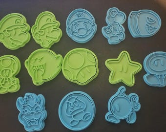 Puede incluir: Un conjunto de cortadores de galletas de plástico verde y azul con personajes de un videojuego popular. El conjunto incluye a Mario, Luigi, Yoshi, un Bill Bala, un hongo, una estrella y otras figuras reconocibles.