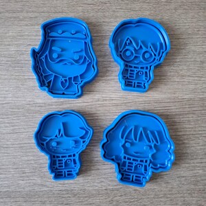 May include: Four blue plastic cookie cutters featuring character designs. The cutters depict a wizard with a beard, a boy with glasses, a boy with messy hair, and a girl with wavy hair. The cutters are arranged on a wooden surface.