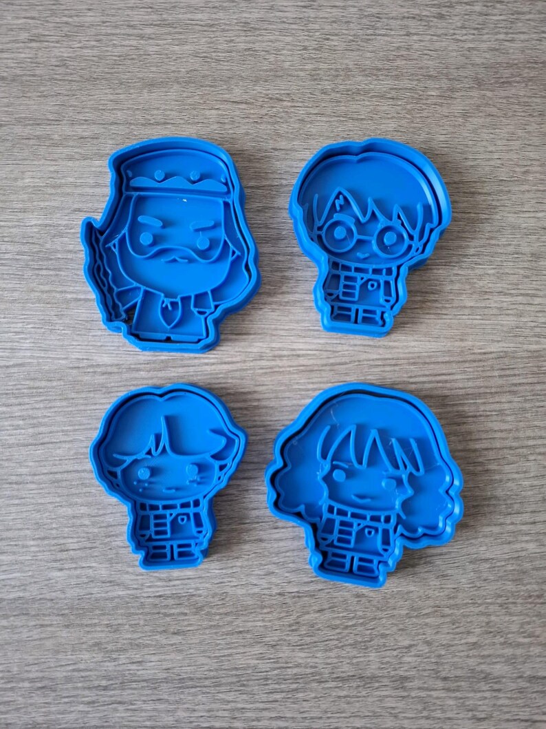May include: Four blue plastic cookie cutters featuring character designs. The cutters depict a wizard with a beard, a boy with glasses, a boy with messy hair, and a girl with wavy hair. The cutters are arranged on a wooden surface.