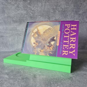 May include: A green book stand holding a copy of "Harry Potter and the Prisoner of Azkaban" by J.K. Rowling. The book cover features a fantasy illustration with the title in purple. The stand is a bright, solid green.