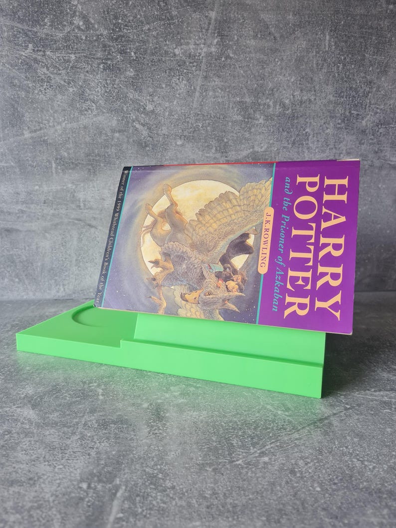 May include: A green book stand holding a copy of "Harry Potter and the Prisoner of Azkaban" by J.K. Rowling. The book cover features a fantasy illustration with the title in purple. The stand is a bright, solid green.