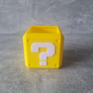 May include: A yellow, cube-shaped container featuring a white question mark on the front. The container has a rectangular opening at the top and small circular details on the sides. The design is inspired by a video game.