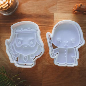 May include: Two white plastic cookie cutters in the shape of cartoon characters. One is a bearded king with a crown, and the other is a figure holding a wand. The cutters are on a wooden surface.