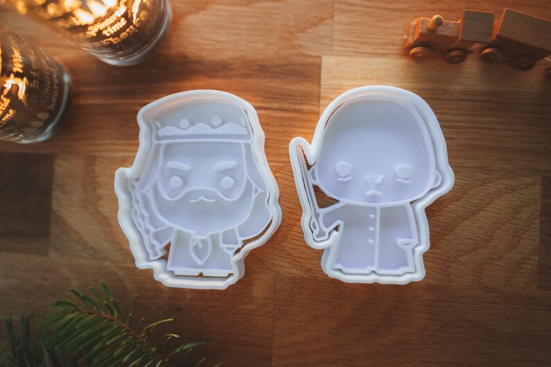May include: Two white plastic cookie cutters in the shape of cartoon characters. One is a bearded king with a crown, and the other is a figure holding a wand. The cutters are on a wooden surface.