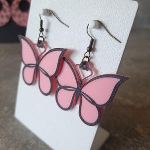 May include: Pink butterfly earrings with dark gray outlines and matching ear hooks. The earrings are displayed on a white card. The butterfly design is visible on the earrings.