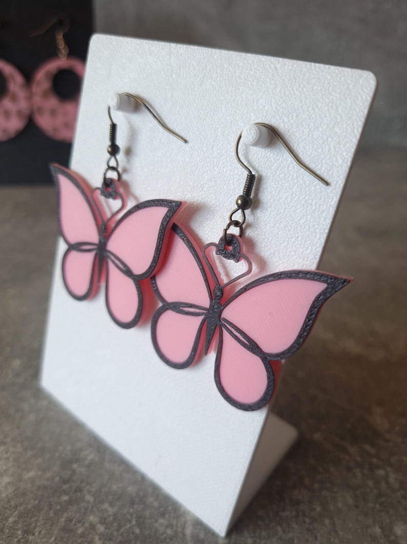 May include: Pink butterfly earrings with dark gray outlines and matching ear hooks. The earrings are displayed on a white card. The butterfly design is visible on the earrings.