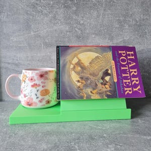 May include: A floral-patterned mug with a pink handle and rim sits on a bright green book stand next to a copy of "Harry Potter and the Prisoner of Azkaban" by J.K. Rowling. The book cover features an illustration of a figure riding a winged creature.