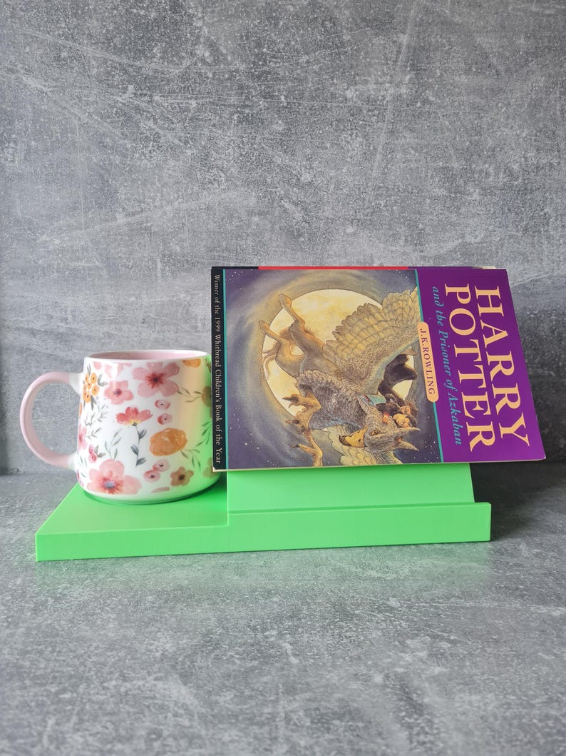 May include: A floral-patterned mug with a pink handle and rim sits on a bright green book stand next to a copy of "Harry Potter and the Prisoner of Azkaban" by J.K. Rowling. The book cover features an illustration of a figure riding a winged creature.