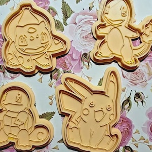May include: Four orange plastic cookie cutters featuring popular cartoon characters. The cutters include Bulbasaur, Charmander, Squirtle, and Pikachu. The cookie cutters are on a floral background.