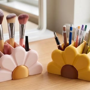 May include: Two flower-shaped desk organizers, one light pink and one mustard yellow, each featuring a central circular accent. The pink organizer holds makeup brushes and cosmetic tubes, while the yellow organizer contains various pens and markers.