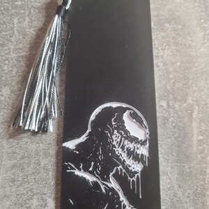 May include: Black bookmark with a white image of Venom, a fictional superhero character, with a black tassel.