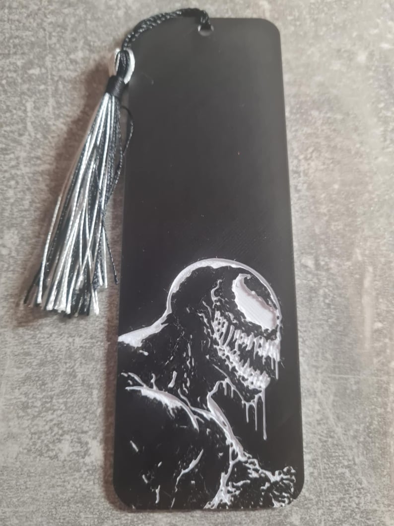 May include: Black bookmark with a white image of Venom, a fictional superhero character, with a black tassel.