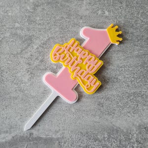 Op de afbeelding: Een roze en gele taarttopper met de tekst "Happy Birthday" en het cijfer "1". De topper heeft een kleine gele kroon en een witte prikker om in een taart te steken. Het ontwerp is gelaagd met een witte omtrek.