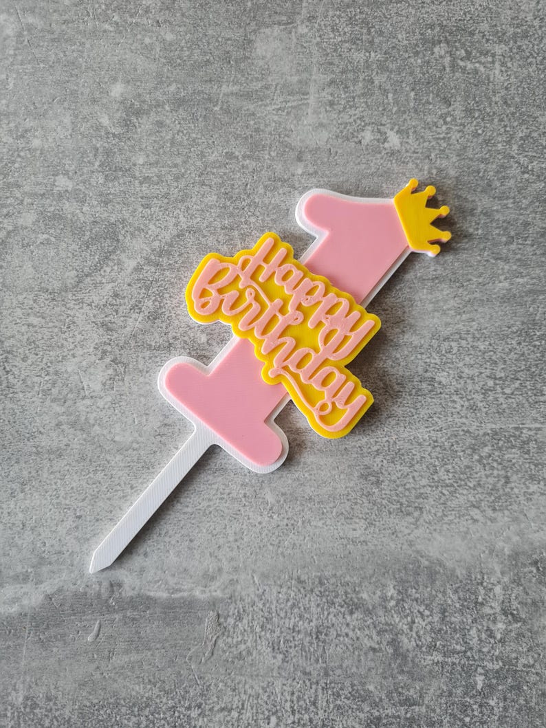 Op de afbeelding: Een roze en gele taarttopper met de tekst "Happy Birthday" en het cijfer "1". De topper heeft een kleine gele kroon en een witte prikker om in een taart te steken. Het ontwerp is gelaagd met een witte omtrek.