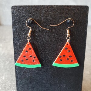 May include: Watermelon slice earrings with red and black details, and a green rind. The earrings are suspended from gold-colored ear wires. The earrings are displayed on a black surface.