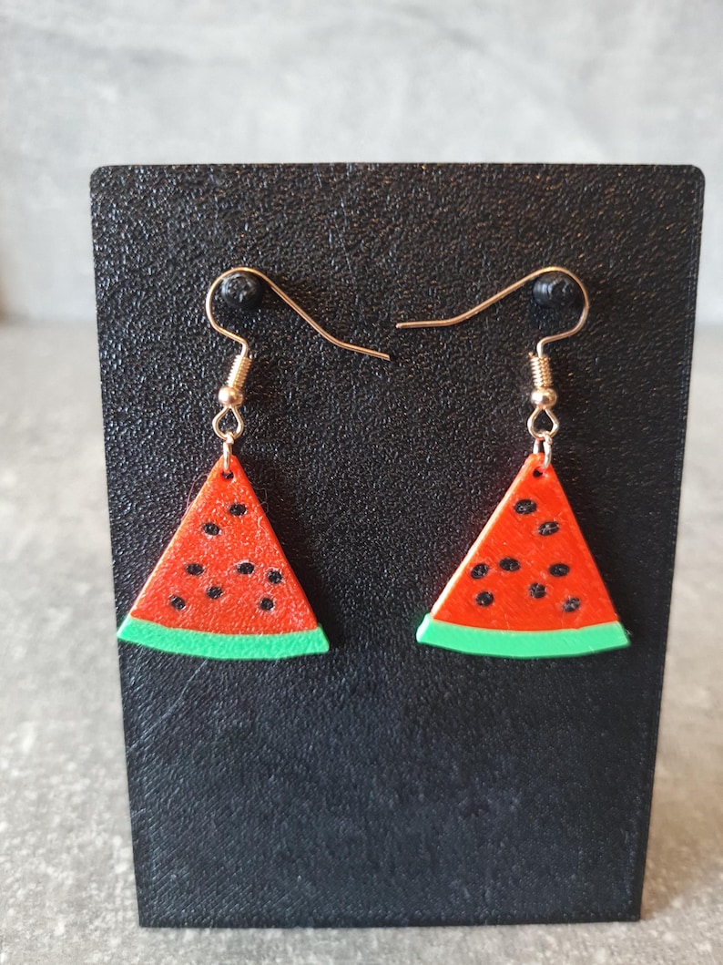 May include: Watermelon slice earrings with red and black details, and a green rind. The earrings are suspended from gold-colored ear wires. The earrings are displayed on a black surface.