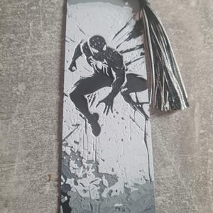 May include: Black and white bookmark featuring a silhouette of Spider-Man in a jumping pose. The bookmark has a gray and white textured background and a black and white tassel.