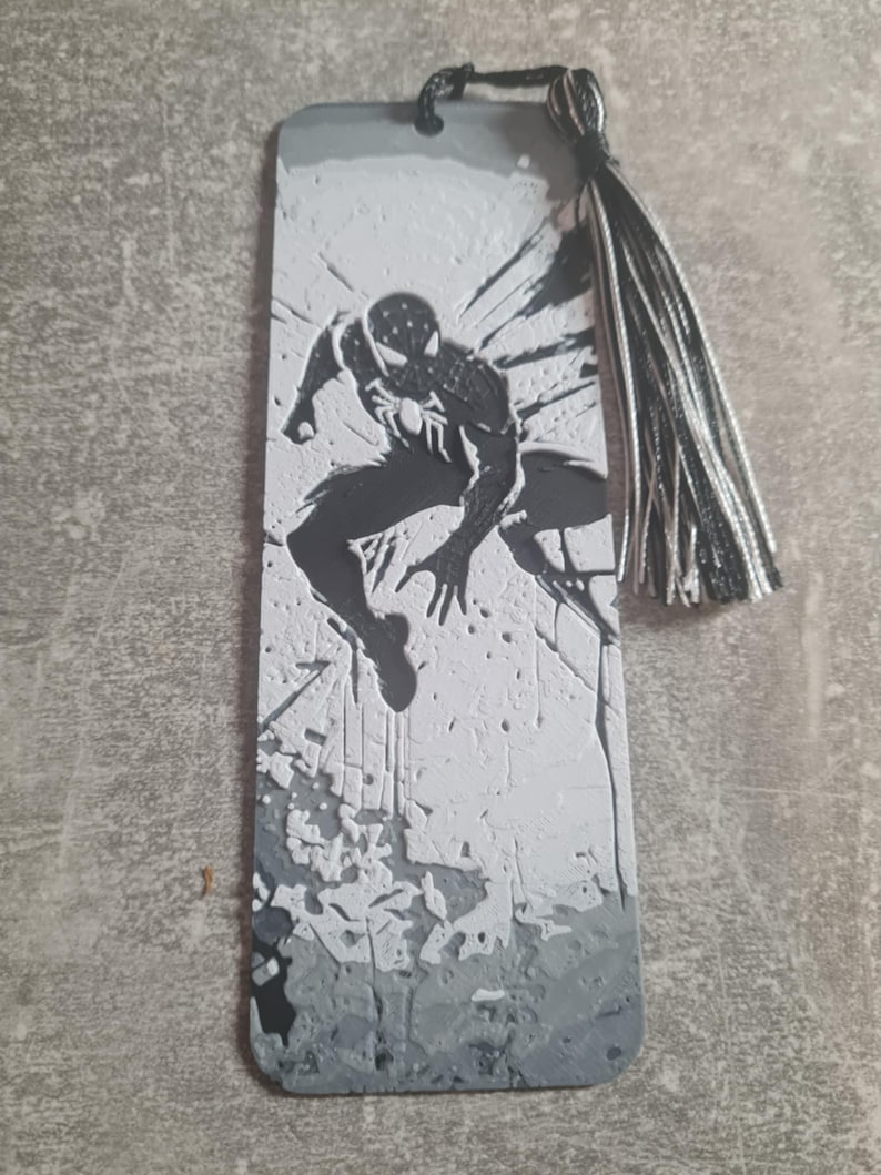 May include: Black and white bookmark featuring a silhouette of Spider-Man in a jumping pose. The bookmark has a gray and white textured background and a black and white tassel.