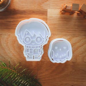 May include: Two white plastic cookie cutters in the shape of a character with glasses and a lightning bolt scar, and a small owl. The cutters are on a wooden surface, with a wooden toy train and greenery in the background.