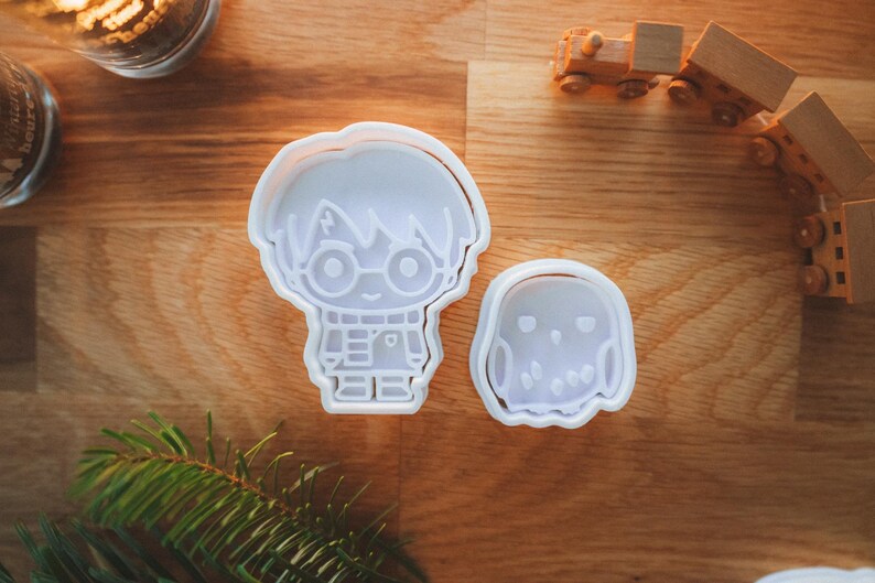 May include: Two white plastic cookie cutters in the shape of a character with glasses and a lightning bolt scar, and a small owl. The cutters are on a wooden surface, with a wooden toy train and greenery in the background.