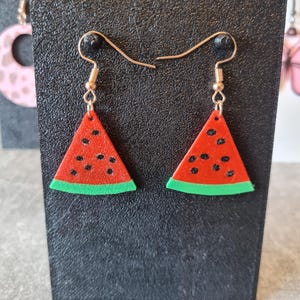 May include: Watermelon slice earrings with gold-colored hooks. The earrings are red with black seeds and a green rind. The earrings are displayed on a black surface.