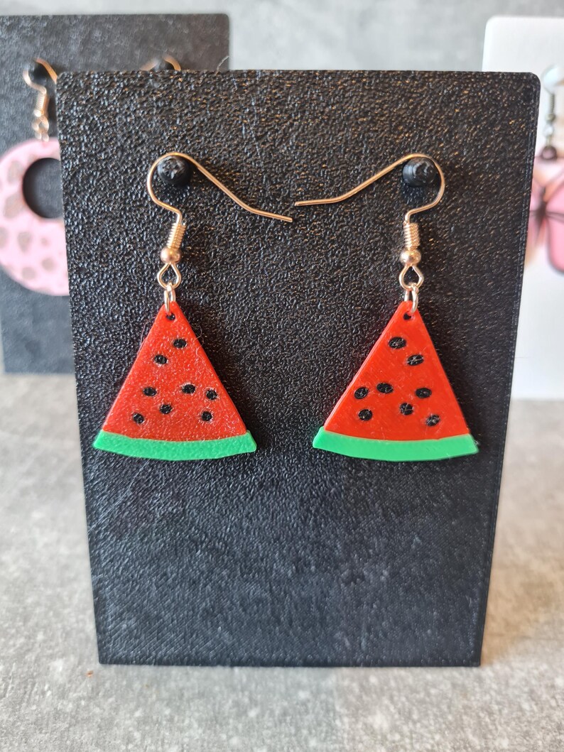 May include: Watermelon slice earrings with gold-colored hooks. The earrings are red with black seeds and a green rind. The earrings are displayed on a black surface.