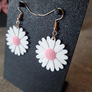 May include: A pair of daisy earrings with white petals and a pink center. The earrings are suspended from gold-colored ear wires. The earrings are displayed on a black textured surface.