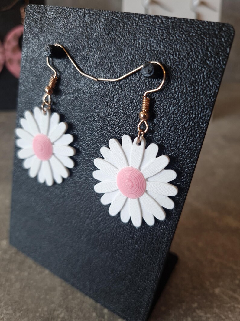 May include: A pair of daisy earrings with white petals and a pink center. The earrings are suspended from gold-colored ear wires. The earrings are displayed on a black textured surface.