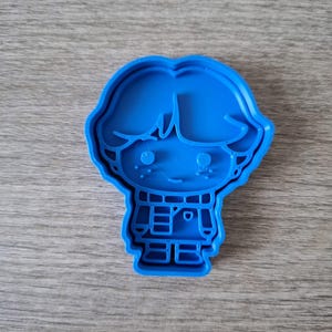May include: A blue plastic cookie cutter in the shape of a cartoon boy. The cutter features a detailed outline of a boy with hair, eyes, a smile, and a collared shirt. The cookie cutter is designed for baking and crafting.