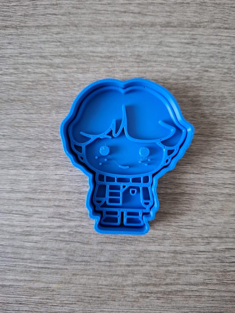May include: A blue plastic cookie cutter in the shape of a cartoon boy. The cutter features a detailed outline of a boy with hair, eyes, a smile, and a collared shirt. The cookie cutter is designed for baking and crafting.