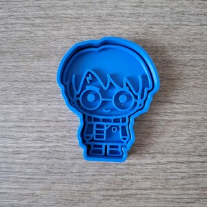 May include: A blue plastic cookie cutter in the shape of a cartoon character with glasses and a scarf. The cutter is on a wooden surface.