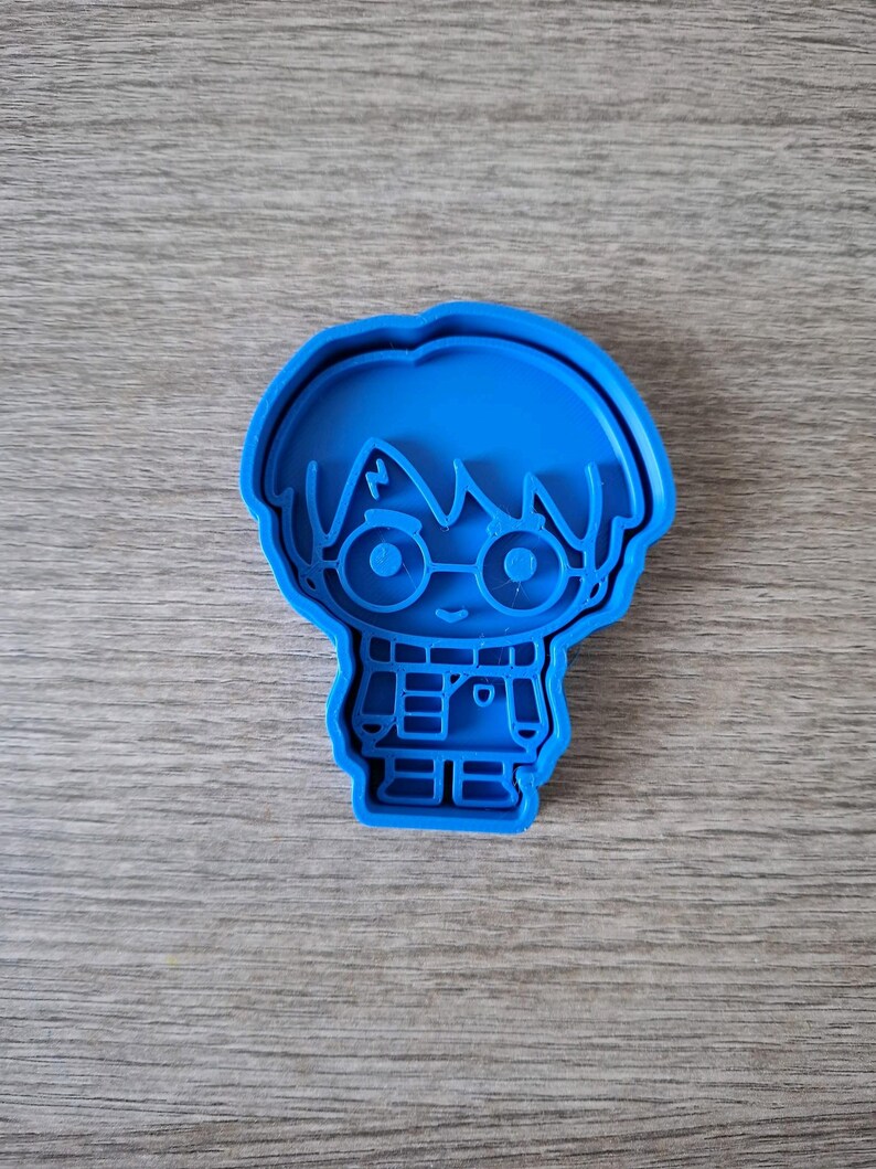 May include: A blue plastic cookie cutter in the shape of a cartoon character with glasses and a scarf. The cutter is on a wooden surface.