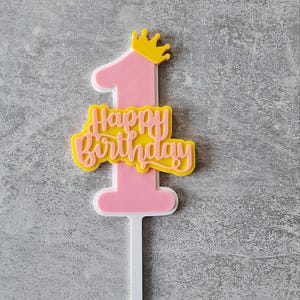 Op de afbeelding: Roze en gele taarttopper met het cijfer "1", een gele kroon en de tekst "Happy Birthday" in sierlijke letters. De topper heeft een witte basis om in de taart te steken. Geschikt voor een eerste verjaardag.