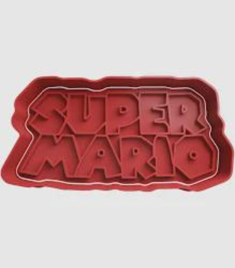 May include: Red plastic cookie cutter in the shape of the words "SUPER MARIO". The letters are raised and the cutter has a rectangular outline. The cutter is designed for making biscuits or other treats.