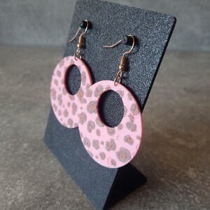 May include: Pink hoop earrings with a brown leopard print pattern. The earrings are displayed on a black stand. The earrings have gold-colored hooks.
