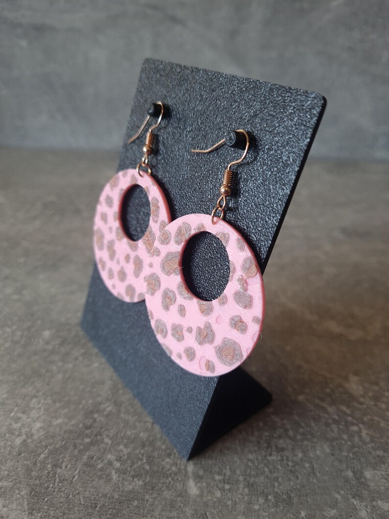 May include: Pink hoop earrings with a brown leopard print pattern. The earrings are displayed on a black stand. The earrings have gold-colored hooks.