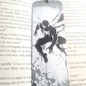 May include: A black and white bookmark featuring a stylized image of Spiderman in a web-like design. The bookmark is made of metal and has a black tassel.