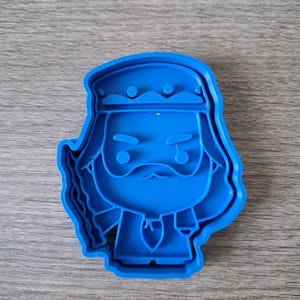 May include: A blue plastic cookie cutter in the shape of a cartoon character with a beard, mustache, and a head covering. The cutter has a detailed outline and is designed for creating cookies or other treats.